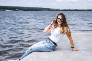Uzun saçlı, şık gözlüklü, gölün yanındaki beton sahilde poz veren genç bir kadın. Kot pantolon ve tişört giymiş gülümseyen ve kameraya bakan bir kız..
