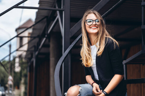 Красивая девушка с длинными волосами и очками сидит на металлической лестнице на деревянном фоне дома с вертикальными досками. Женщина улыбается и смотрит в камеру
.