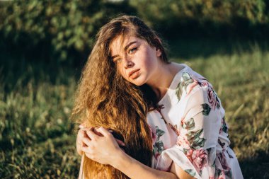 Uzun kıvırcık saçlı, esmer, çilli, makyajlı, yeşil gözlü, kamera önünde poz veren açık beyaz elbiseli, arka planı bulanık, doğa...