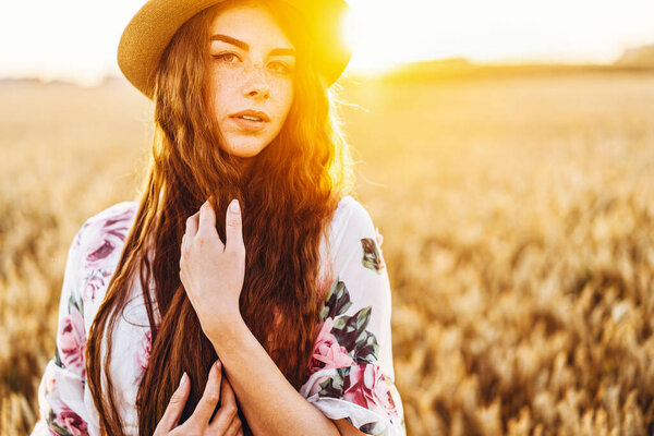 Portrait of a beautiful young woman with curly hair and freckles face. Woman in dress and hat posing in wheat field at sunset and looking at camera.