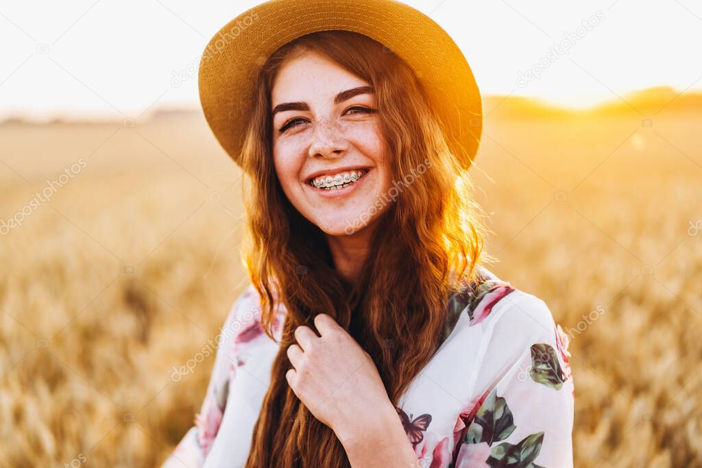 Portrait of a beautiful young woman with curly hair and freckles face. Woman in dress and hat posing in wheat field at sunset and looking at camera.