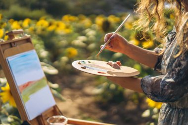 Close up hands of female artist holding brush and palette with oil paints. Blurred background with easel in sunflower field. clipart