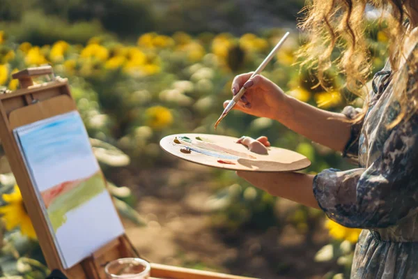 Close up hands of female artist holding brush and palette with oil paints. Blurred background with easel in sunflower field.