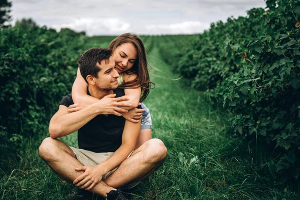 A man sitting on green grass between currant bushes, a woman with long hair hugging him from behind. Background of currant plantations.