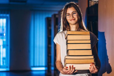 Kütüphanede gözlüklü genç bir kız gülümsüyor ve bir yığın kitap taşıyor. Sınav hazırlığı.
