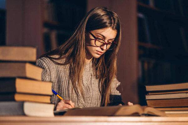Young student in glasses preparing for the exam. Girl in the evening sits at a table in the library with a pile of books.