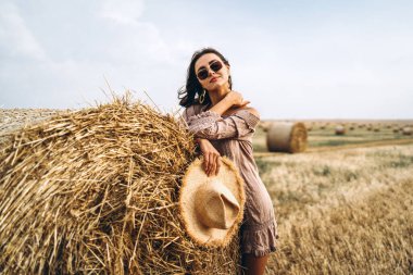 Buğday tarlasında ve saman balyalarında çıplak omuzlu, güneş gözlüklü gülümseyen kadın..