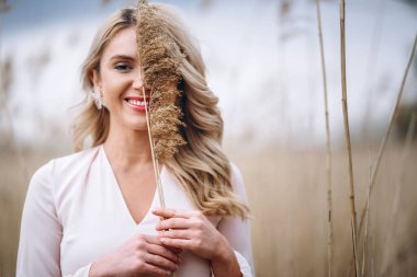 Uzun sarı kıvırcık saçlı, güzel bir kızın fotoğrafı. Sazlık tarlasında duruyor ve elinde yüksek kamış dalları var.