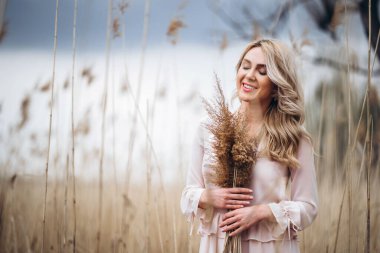 Uzun sarı kıvırcık saçlı, güzel bir kızın fotoğrafı. Sazlık tarlasında duruyor ve elinde yüksek kamış dalları var.