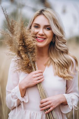 Uzun sarı kıvırcık saçlı, güzel bir kızın fotoğrafı. Sazlık tarlasında duruyor ve elinde yüksek kamış dalları var.