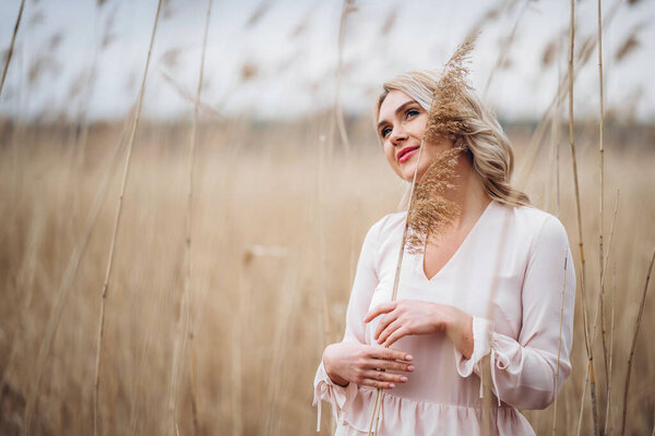 Photo of a pretty smiling girl with long blond curly hair in light long drees standing in a reed field and holding high reed branches in her hands
