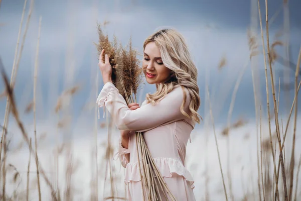 照片上是一个漂亮的微笑着的女孩 一头长长的金发卷曲在淡淡的长长的芦苇里 站在芦苇地里 手里拿着高高的芦苇枝条 — 图库照片