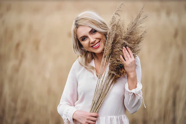 照片上是一个漂亮的微笑着的女孩 一头长长的金发卷曲在淡淡的长长的芦苇里 站在芦苇地里 手里拿着高高的芦苇枝条 — 图库照片