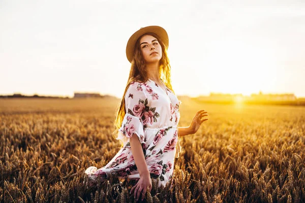 一个有着卷发和雀斑的漂亮年轻女子的画像 日落时分 身穿连衣裙 头戴帽子的女人站在麦田里 看着镜头 — 图库照片