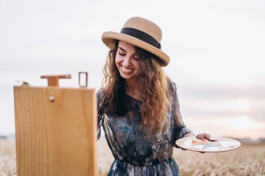 Şapkalı, kıvırcık saçlı, gülümseyen kadın sanatçının portresi. Kız buğday tarlasında bir manzara resmi çiziyor.. 