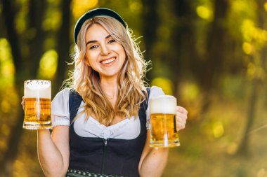 Dirndl giymiş mutlu bir sarışın, geleneksel festival elbisesi, arka planda bulanık bir şekilde ormanda bir kupa bira tutuyor. Ekim Festivali, uluslararası bira günü konsepti