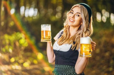 Dirndl giymiş mutlu bir sarışın, geleneksel festival elbisesi, arka planda bulanık bir şekilde ormanda bir kupa bira tutuyor. Ekim Festivali, uluslararası bira günü konsepti