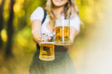 Dirndl giymiş mutlu bir sarışın, geleneksel festival elbisesi, arka planda bulanık bir şekilde ormanda bir kupa bira tutuyor. Ekim Festivali, uluslararası bira günü konsepti