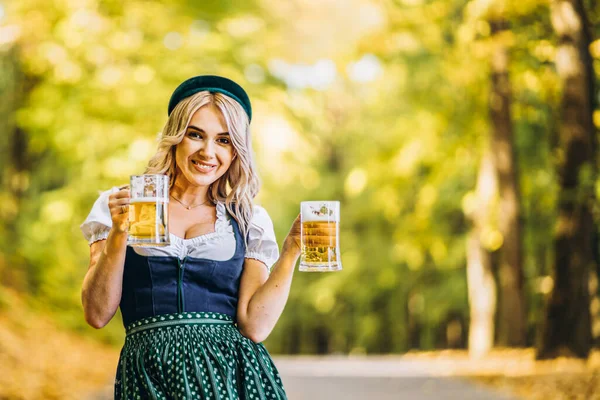 汚れでかなり幸せなブロンド 伝統的な祭りのドレス ぼやけた背景と森の中で屋外ビールのマグカップを保持 オクトーバーフェスト 国際的なビールの日のコンセプト — ストック写真