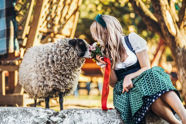 ブロンド女の子で伝統的なOktoberfestドレスで花束の花の近くに座っているかなり羊上の農場 — ストック写真