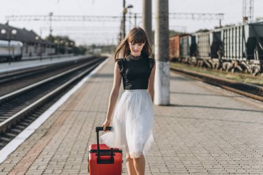 Beyaz etekli, kırmızı valizli, trenini bekleyen ve tren istasyonunda yürüyen esmer bir kadın yolcu.