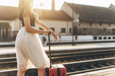 Beyaz etekli, kırmızı valizli, trenini bekleyen ve tren istasyonunda yürüyen esmer bir kadın yolcu.