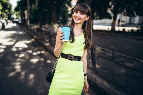 在城市的大街上 穿着浅绿色衣服 头戴蓝色纸杯饮料的漂亮的黑发女孩走在大街上 — 图库照片
