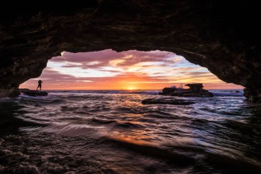 Mağaranın içinden görünen görkemli bir gün doğumu. Uzakta bir adamın silueti var. Bu Avustralya, Yeni Güney Galler 'deki Cave Beach' te çekildi.