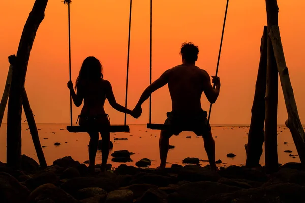 Silhouette of a couple holding hands while sitting on big swings at sunset