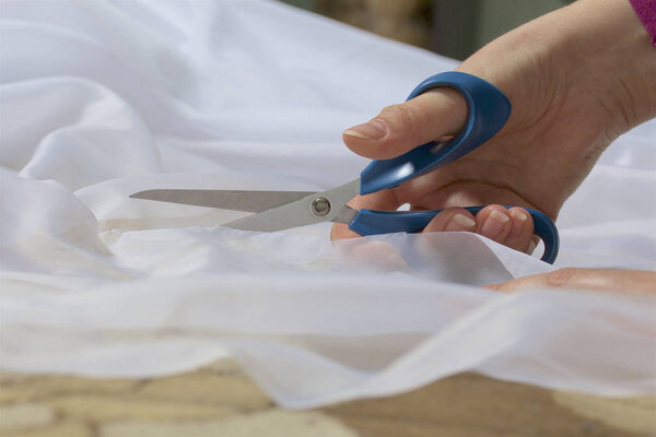 The woman cuts the fabric with scissors for sewing curtains on the window.