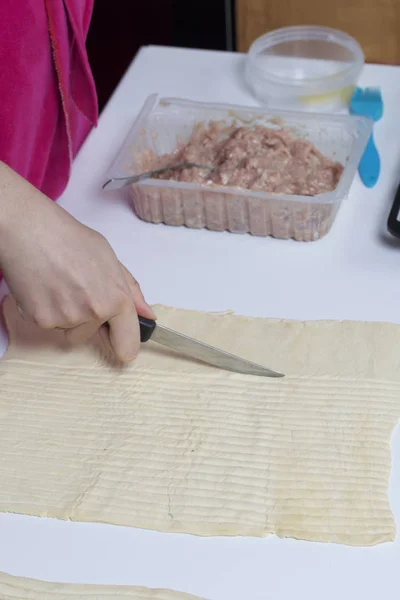 Stages of preparation of meat glomeruli. A woman slices thin strips of dough. Next to the table is a stuffing and lie tools.