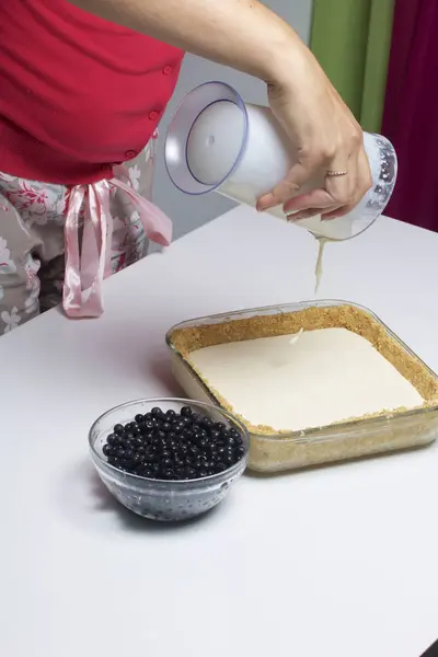 The woman adds the filling to the biscuit.  Delicate cheesecake with blueberries. Ingredients: cookies and lemon stand on the table.