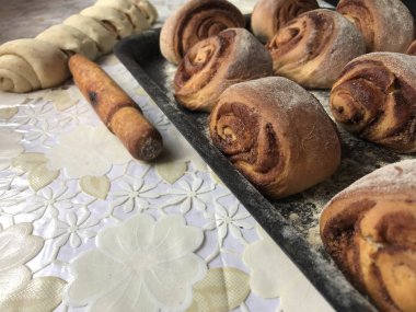 Evde tarçın ile buns hazırlanması. Hazır buns fırın tepsisine yalan. Tablo yanında bazı ham demir çubuklar ve ahşap bir oklava vardır.