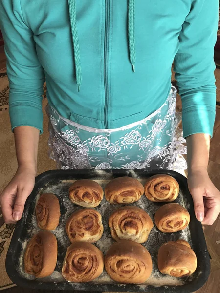 Preparação de pães com canela em casa. Uma mulher em um avental segura uma bandeja de cozimento com rolos de pão . — Fotografia de Stock