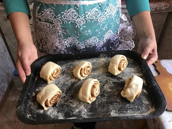 Evde tarçın ile buns hazırlanması. Bir kadın apronda fırın tepsisi topuz rulo ile taşıyabilir.