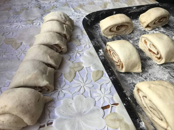 Preparation of buns with cinnamon at home. Raw buns lie on a baking sheet. Next to the table are some raw billets and a wooden rolling pin. — Stock Photo, Image