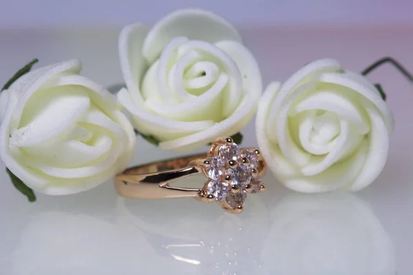 Διακοσμητικά για τους εορτασμούς. Χρυσό δαχτυλίδι με πέτρες και μπουκέτο των τεχνητών λουλουδιών. Τριαντάφυλλα μπεζ σε λευκό φόντο. — Φωτογραφία Αρχείου