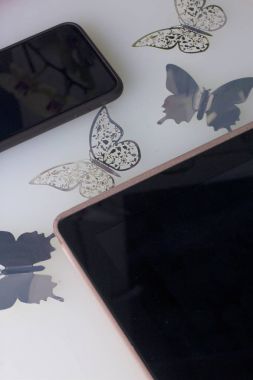 Beyaz bir yüzeyde akıllı telefon ve tablet bulunmaktadır. Çevresinde folyo kesilmiş kelebekler yapılmış süsler.