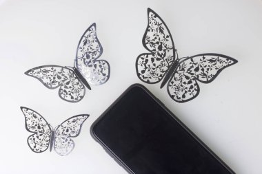 Akıllı telefon beyaz bir yüzeyde duruyor. Onun etrafında kelebekler süsler, folyo kesilmiş.
