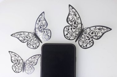 Akıllı telefon beyaz bir yüzeyde duruyor. Onun etrafında kelebekler süsler, folyo kesilmiş.