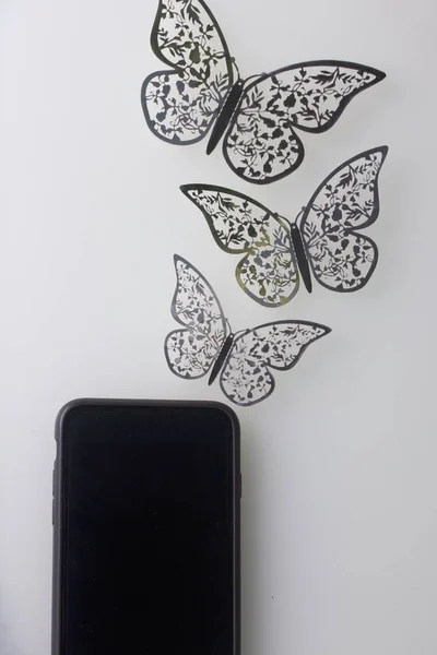 Smartphone se opírá o bílý povrch. Kolem něho ozdoby motýlů, řezané z fólie. — Stock fotografie