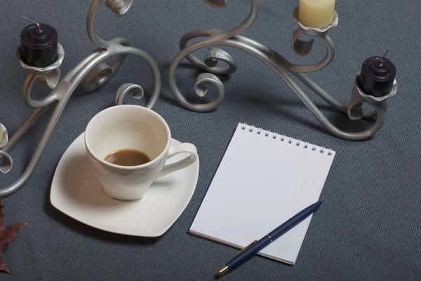 Bougeoir en métal forgé avec bougies. Une tasse avec du café non approuvé. Il y a un bloc-notes ouvert et un stylo. Les feuilles tombées en automne de jaune et de rouge sont éparpillées à la surface . — Photo