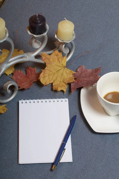 Bougeoir en métal forgé avec bougies. Une tasse avec du café non approuvé. Il y a un bloc-notes ouvert et un stylo. Les feuilles tombées en automne de jaune et de rouge sont éparpillées à la surface . — Photo