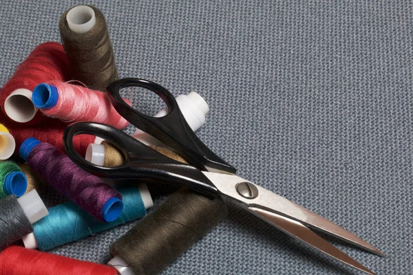 Cívky nití různých barev na pozadí šedé tkaniny a nůžky. — Stock fotografie