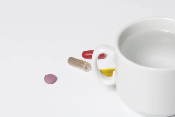 En la superficie blanca de la mesa se encuentran píldoras de diferentes colores y hay una taza con agua . —  Fotos de Stock