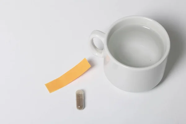 En la superficie blanca de la mesa hay una pastilla y una taza con agua. Siguiente es una pegatina de papel con un recordatorio . —  Fotos de Stock