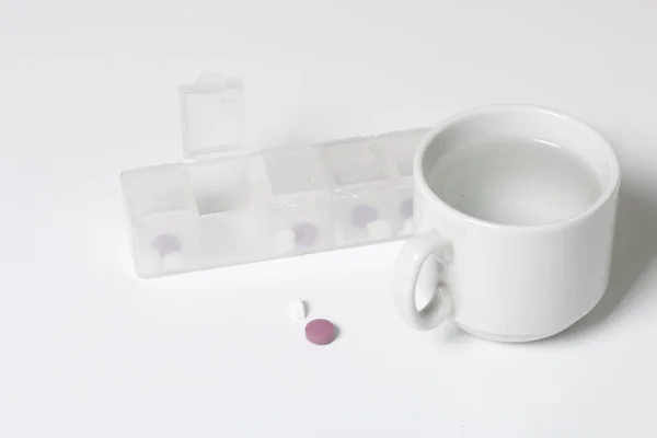 Планшет з таблетками різних кольорів. Одна клітина відкрита, таблетки від неї лежать поруч на столі. Поруч є кухоль з водою. . — стокове фото