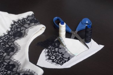 Dikiş ve sos. Karanlık yüzeyde dekorasyon için dikilmiş bir siyah kurdele ile beyaz bir elbise var. Yakınlarda makas, iplik makaraları yalan ve ürünün malzeme süsleme.