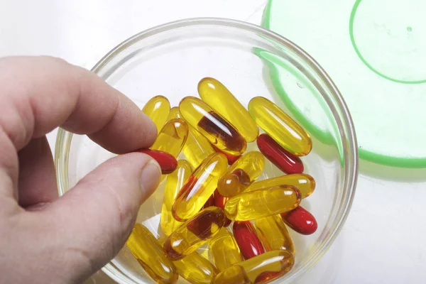 El hombre tiene una pastilla en la mano. Las píldoras de color rojo y amarillo están en un recipiente de vidrio con una tapa de plástico. Sobre una superficie blanca. Vista desde arriba . —  Fotos de Stock