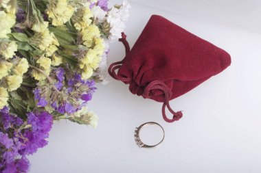 Kurutulmuş çiçek bir buket beyaz bir yüzeye yatıyor. Sırada bir hediye ile bir kadife çanta var. Sevgiliniz için yüzük.
